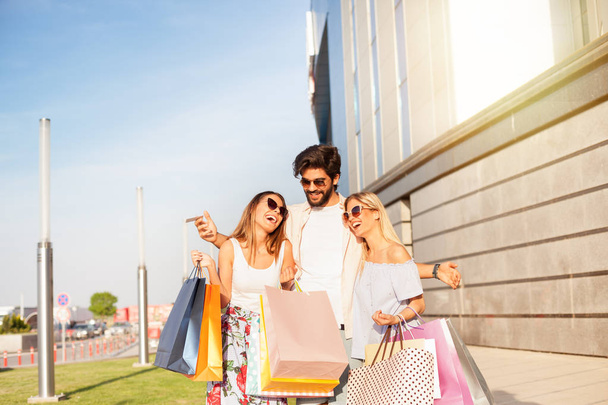 Rich young man with two girls and lot of colored shopping bags and laughing in mall, concept of consumerism, sale, rich life. - Photo, image