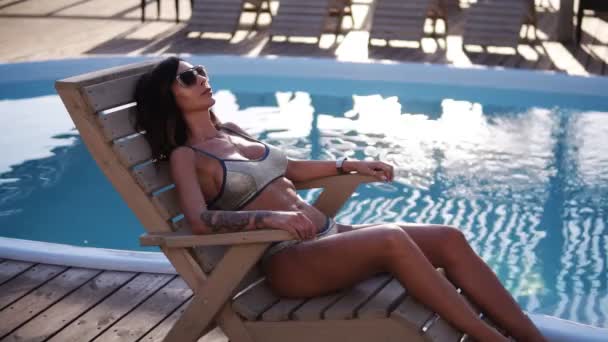 A young woman with a stunning, fit body sunbathes on a trestle by the pool. A girl in a stylish fashion swimsuit and sunglasses. The sun shines on the water - Video, Çekim