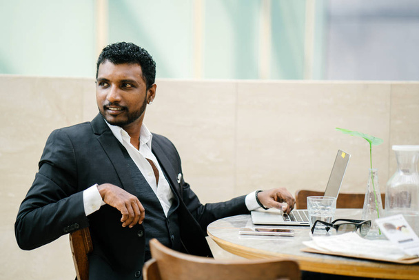 handsome and roguish Indian Asian man in a 3 piece suit is working on his laptop. He is seated at a table during the day and looks calm - Fotoğraf, Görsel