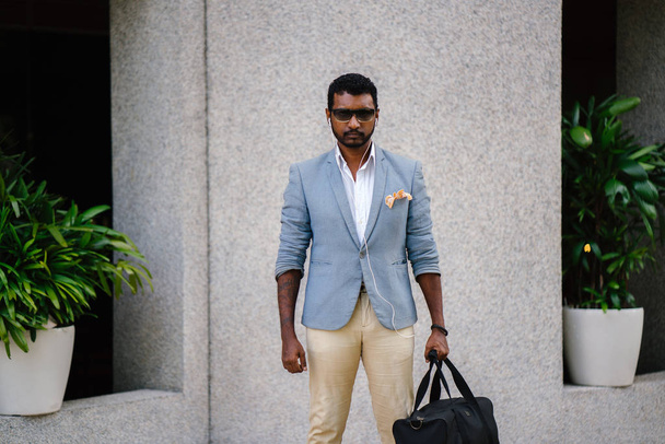 Portrait of a cool Indian Asian man leaning against a wall in the day. He is in a casual suit and sunglasses. He is holding a gym bag as he is lounging in the sun and waiting for someone. - Φωτογραφία, εικόνα