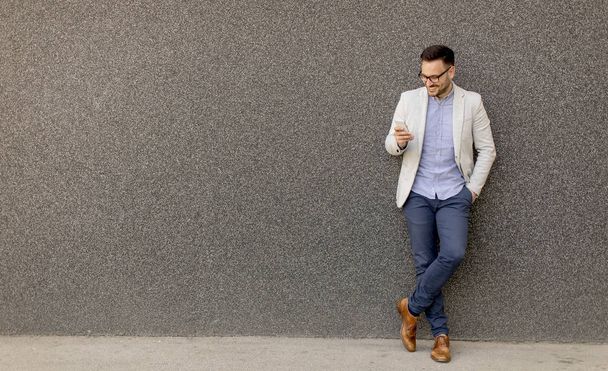 Jonge zakenman in formele slijtage gebruik mobiele telefoon buiten door muur - Foto, afbeelding