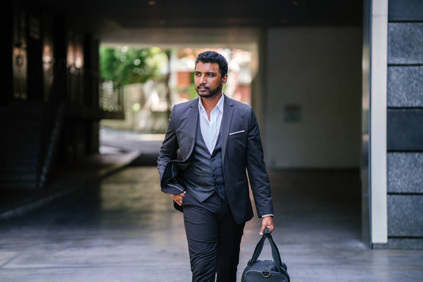 Portrait of a handsome Indian Asian man in a 3-piece suit and holding a bag walking down the street. He is silhouetted against the city and is walking confidently. - Фото, изображение