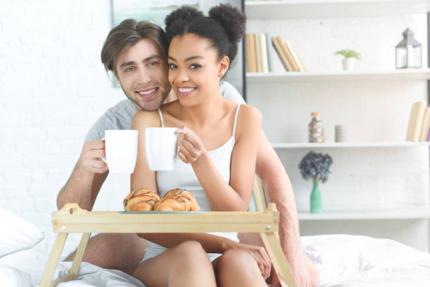 retrato de pareja joven multirracial con tazas de café desayunando en la cama por la mañana en casa
 - Foto, Imagen