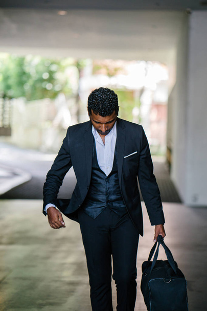 Portrait of a handsome Indian Asian man in a 3-piece suit and holding a bag walking down the street. He is silhouetted against the city and is walking confidently. - Foto, Imagem