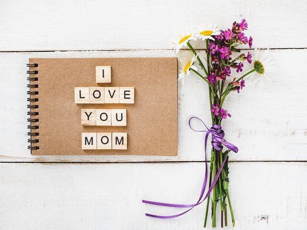 Notepad with wooden letters of the alphabet and the word MOM, a bouquet of wildflowers on a white, vintage, wooden table. Top view, close-up. The concept of a happy family - Zdjęcie, obraz