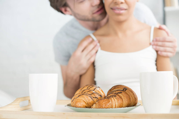 selective focus of breakfast on wooden tray and multiethnic couple at home - Foto, afbeelding