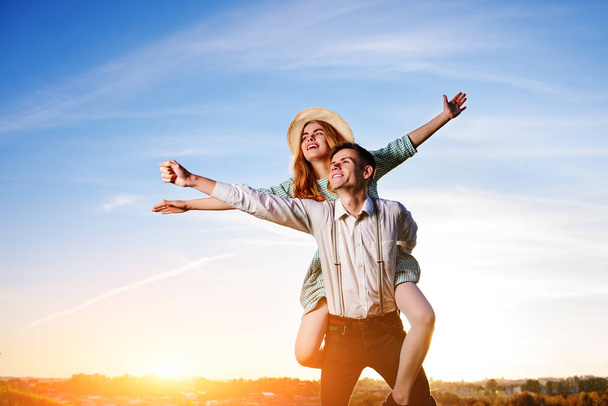 Young guy piggybacking cheerful girlfriend like airplane on the background of sky. - Photo, Image