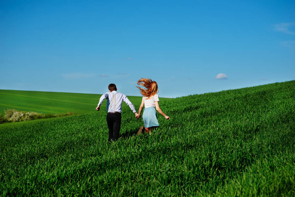Young happy lovers running on meadow with green grass and blue sky, From the back. - Foto, afbeelding