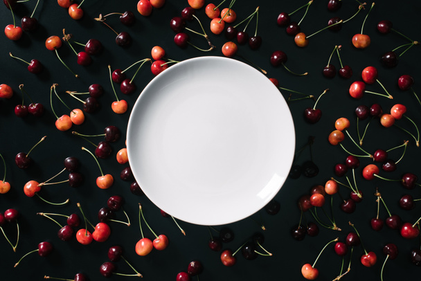 top view of empty round white plate and sweet ripe cherries on black background - 写真・画像