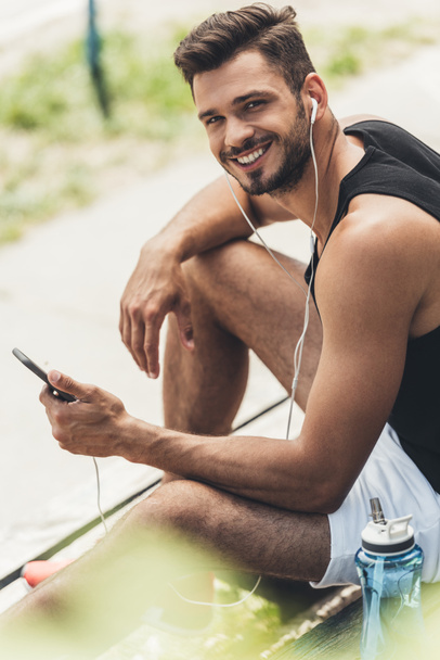 young smiling man with bottle of water listening music with smartphone and earphones on bench  - Fotó, kép