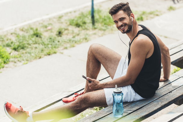 Junger lächelnder Mann mit Wasserflasche hört Musik mit Smartphone und Kopfhörer auf Bank  - Foto, Bild