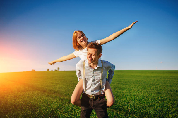 Улыбающийся мужчина держит за спину счастливую женщину, которая вытягивает руки и имитирует полет на фоне голубого неба и зеленого поля
. - Фото, изображение