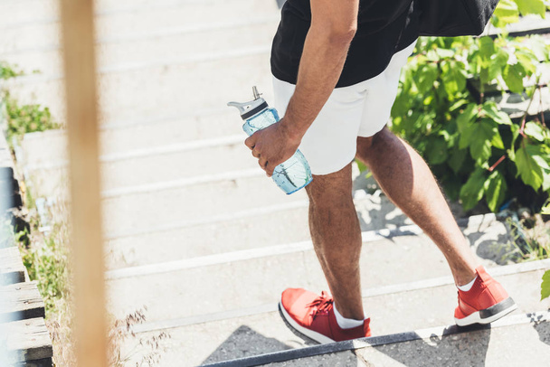 cropped image of sportsman holding bottle of water on stairs at sport playground  - Zdjęcie, obraz
