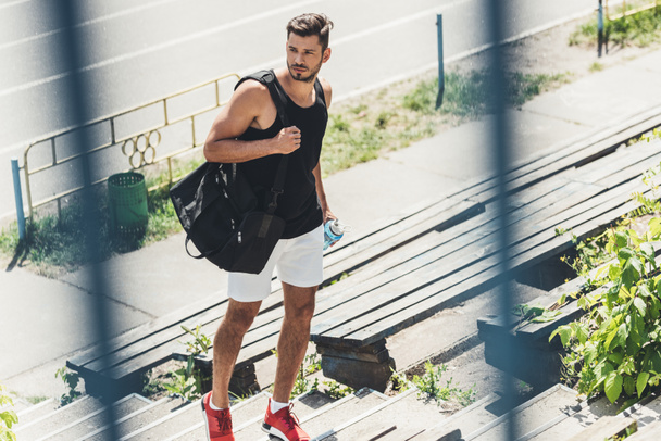 Junger Sportler hält Wasserflasche und Tasche für Sportgeräte auf Treppe am Sportplatz  - Foto, Bild