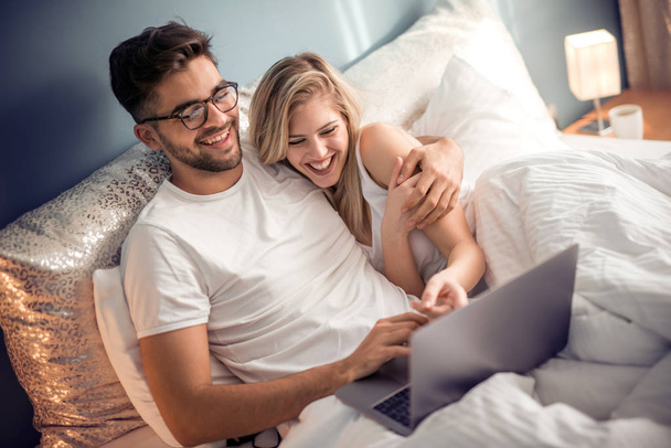 Młoda para z laptopa na łóżku. Mężczyzna i Kobieta, ciesząc się, oglądając film na laptopie podczas relaksu w sypialni. - Zdjęcie, obraz