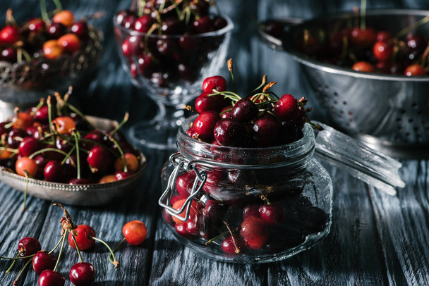 close-up view of fresh ripe cherries in glass jar, colander and utensils on rustic wooden table - Foto, imagen