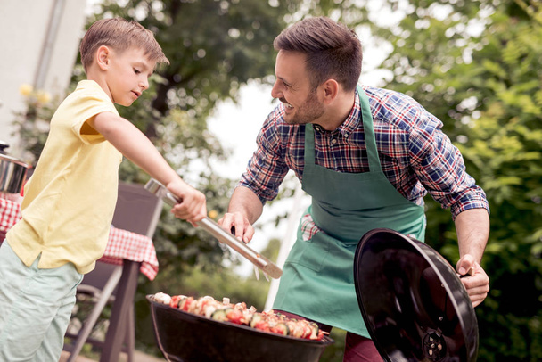 Vrije tijd, voedsel, mensen en feestdagen concept-man koken vlees op de barbecue grill met zijn zoon. - Foto, afbeelding