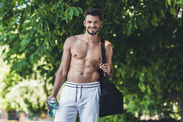 sonriente joven con bolsa para equipo deportivo y botella de fitness mirando a la cámara
 - Foto, imagen