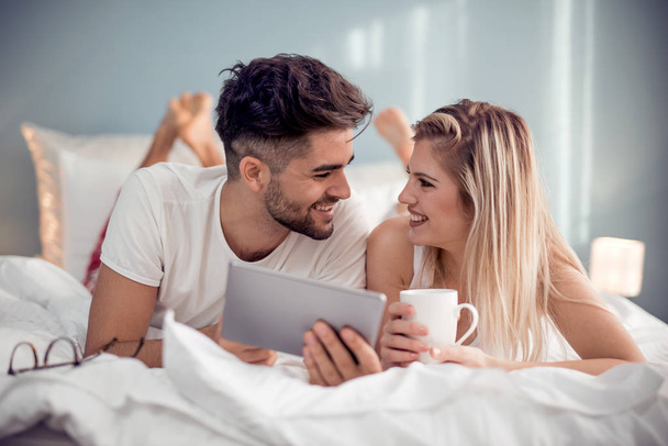 Happy couple in love using tablet in pajamas at home and drink coffee  in bedroom. - Фото, изображение