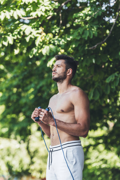 concentrated shirtless young man working out with jumping rope in front of green leaves - Φωτογραφία, εικόνα
