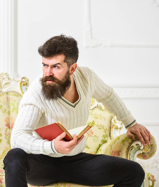 Man with beard and mustache sits on baroque style sofa, holds book, white wall background. Reflections on literature concept. Macho on strict face finished reading book. Guy thinking about literature - Photo, image