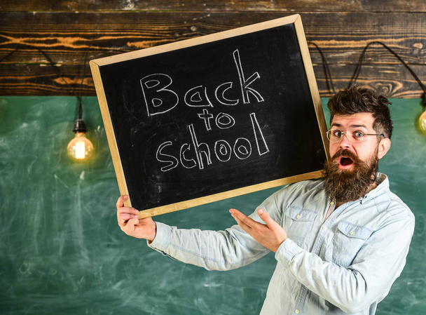 Teacher in eyeglasses holds blackboard with title back to school. Man with beard and mustache on surprised face welcomes students, chalkboard on background. High school concept - Фото, зображення