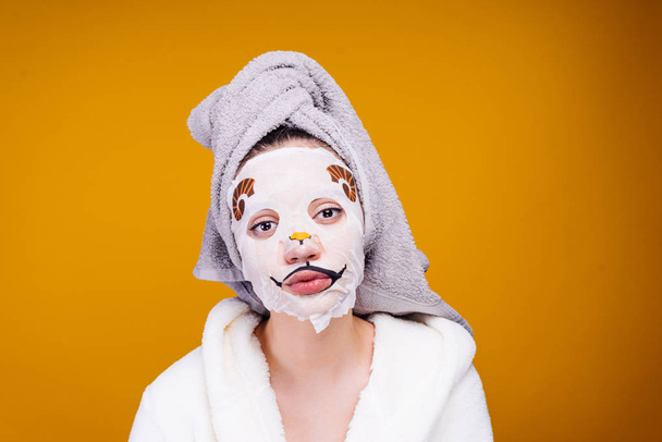 A young girl with a towel on her head and a bathrobe in white clothes takes care of herself, on her face a moisturizing mask - Photo, image