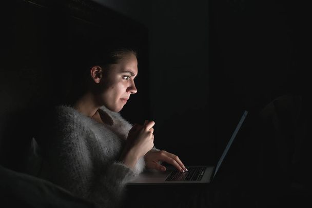 serious young girl sitting in the dark at night on the bed and watching a movie on the laptop - Fotoğraf, Görsel