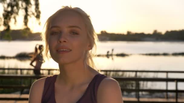 Portrait of young blonde woman looking in camera and smiling, woman running on back, sunset, river and nature background - Video