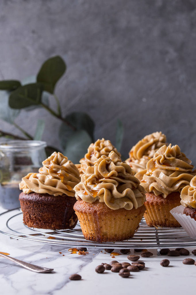 Fresh baked homemade cupcakes with coffee buttercream and caramel standing on cooling rack with eucalyptus branch and coffee beans above over white marble kitchen table. - Foto, Bild