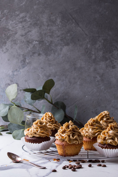 Fresh baked homemade cupcakes with coffee buttercream and caramel standing on cooling rack with eucalyptus branch and coffee beans above over white marble kitchen table. - Foto, Imagem