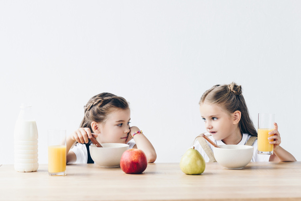 sad schoolgirls eating cereals with orange juice and fruits and looking at each other isolated on white - Photo, Image