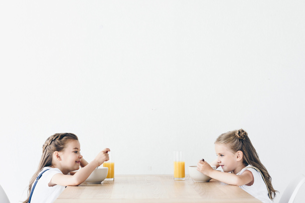 adorable smiling schoolgirls eating cereals with orange juice for breakfast while sitting in front of each other isolated on white - Photo, Image