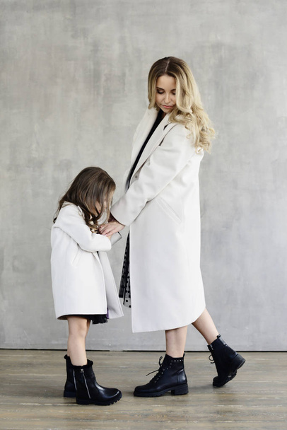 Mom and daughter in warm same white coats holding hands while standing on concrete wall background, lookbook concept - Valokuva, kuva
