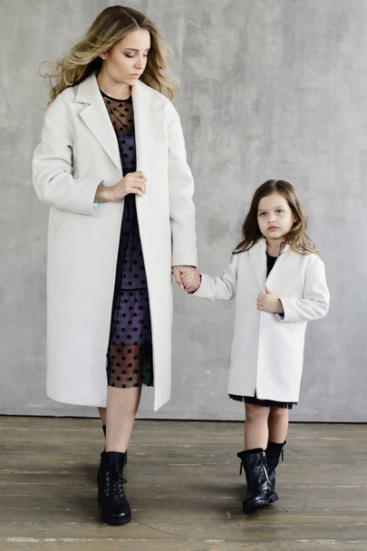 Mutter und Tochter in warmen weißen Mänteln halten Händchen, während sie auf Betonwänden stehen, Lookbook-Konzept - Foto, Bild