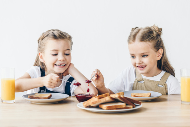 прелестные маленькие сестры едят тосты с джемом на завтрак изолированы на белом
 - Фото, изображение