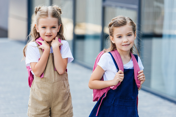 adorable little schoolgirls with pink backpacks walking on street - Zdjęcie, obraz