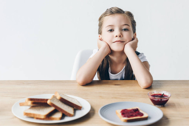 thoughtful little schoolgirl with toasts and jam for breakfast looking at camera isolated on white - 写真・画像