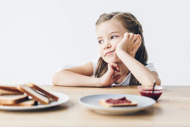 thoughtful little schoolgirl with toasts and jam for breakfast looking away isolated on white - 写真・画像