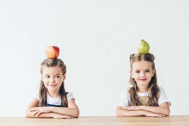 adorable little schoolgirls with apple and pear on heads sitting at table isolated on white - Zdjęcie, obraz
