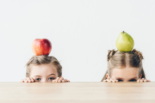 μικρές μαθήτριες με μήλο και αχλάδι στα κεφάλια που κρύβονται πίσω από το τραπέζι που απομονώνονται σε λευκό - Φωτογραφία, εικόνα