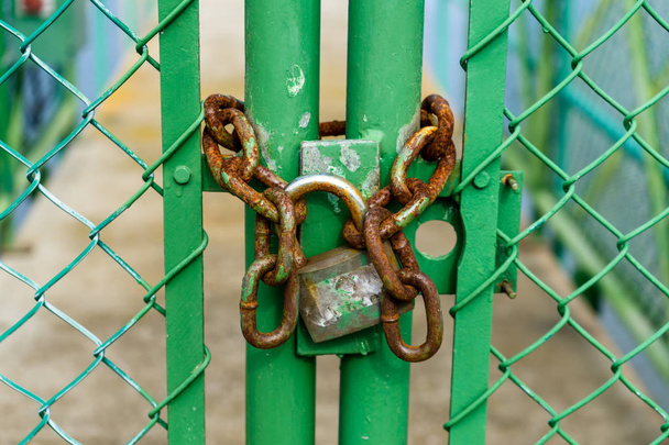 Candado viejo y oxidado encadenado a una puerta verde en una valla que restringe el acceso a un pequeño puente
 - Foto, Imagen