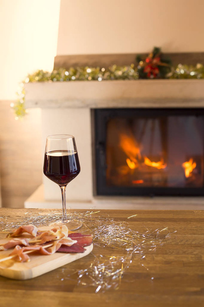 Chimenea decorada para Navidad. Copa de vino tinto y aperitivo en mesa de madera y chimenea para el fondo
. - Foto, imagen