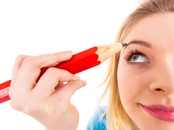 Funny silly woman trying to paint her eyebrows using big huge oversized regular student pencil. Make up fun concept. - 写真・画像