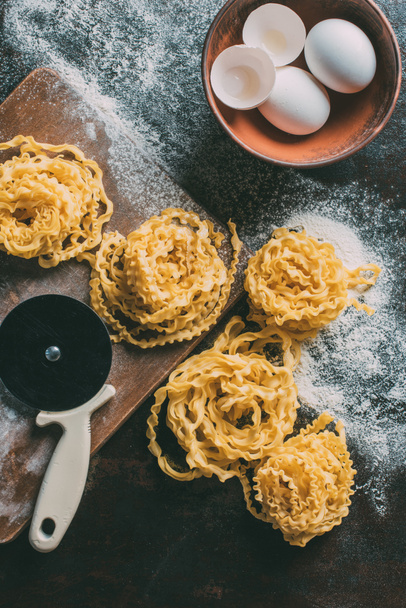 vue du dessus de coupe-pâte réglable, pâtes crues, planche à découper, bol avec coquille d'oeuf et oeufs sur la table couverte de farine
  - Photo, image