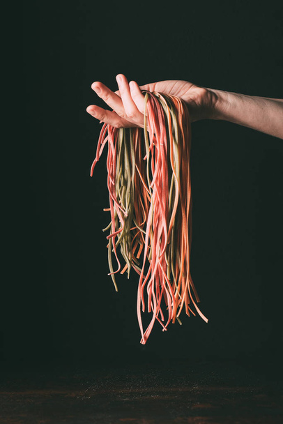 cropped image of man holding homemade raw colorful tagliatelle on hand over table on black background  - Foto, Imagen