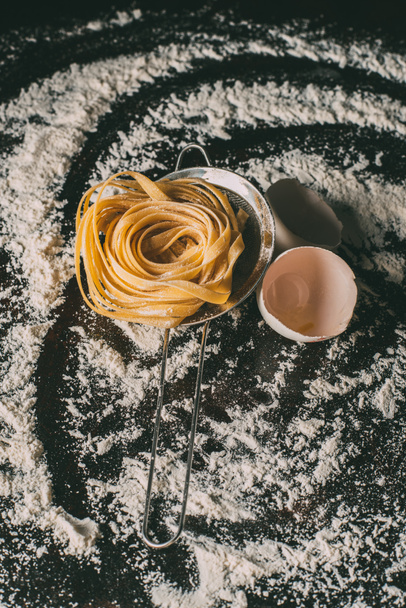 close up image of raw tagliatelle pasta, sieve and egg shells on table covered by flour  - Фото, изображение