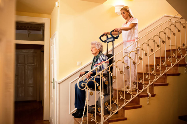 Nurse using machine for climbing to the stairs and helps old mature senior disabled woman at nursing home, care concept - Photo, Image