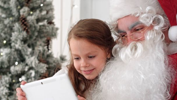 Santa Claus and cute little girl taking Christmas selfies on tablet - Фото, изображение