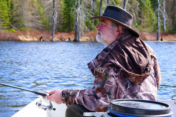   平和な白人男 50 年代、白ひげ、革帽子オーストラリア スタイルと迷彩コートを着ては青い水の湖の彼のボートで釣りの北ケベック州の美しい、新鮮な日当たりの良い朝,                               - 写真・画像